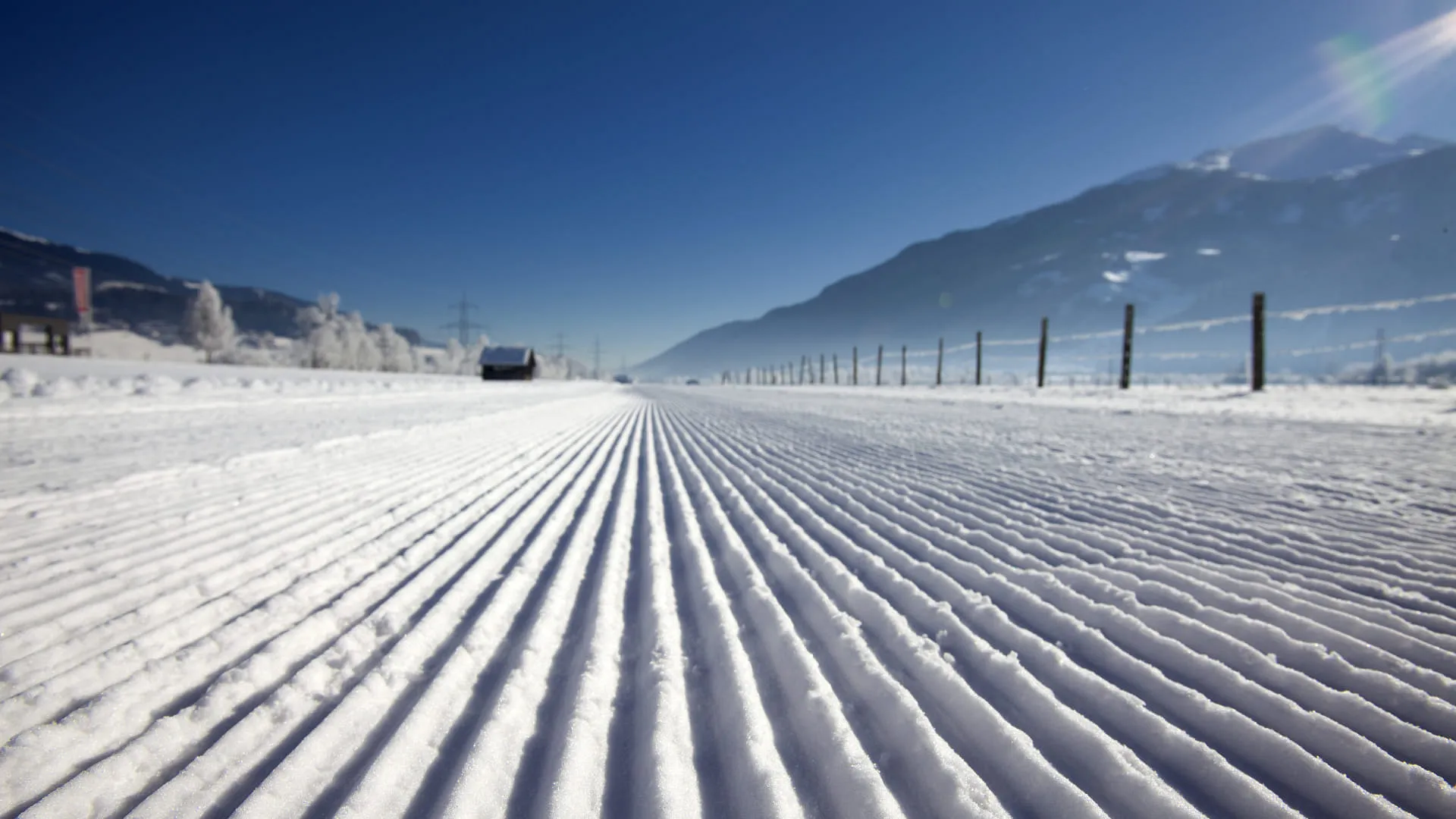 Schneebedeckte Landschaft mit parallel gezogenen Spuren im Schnee und Bergen im Hintergrund.
