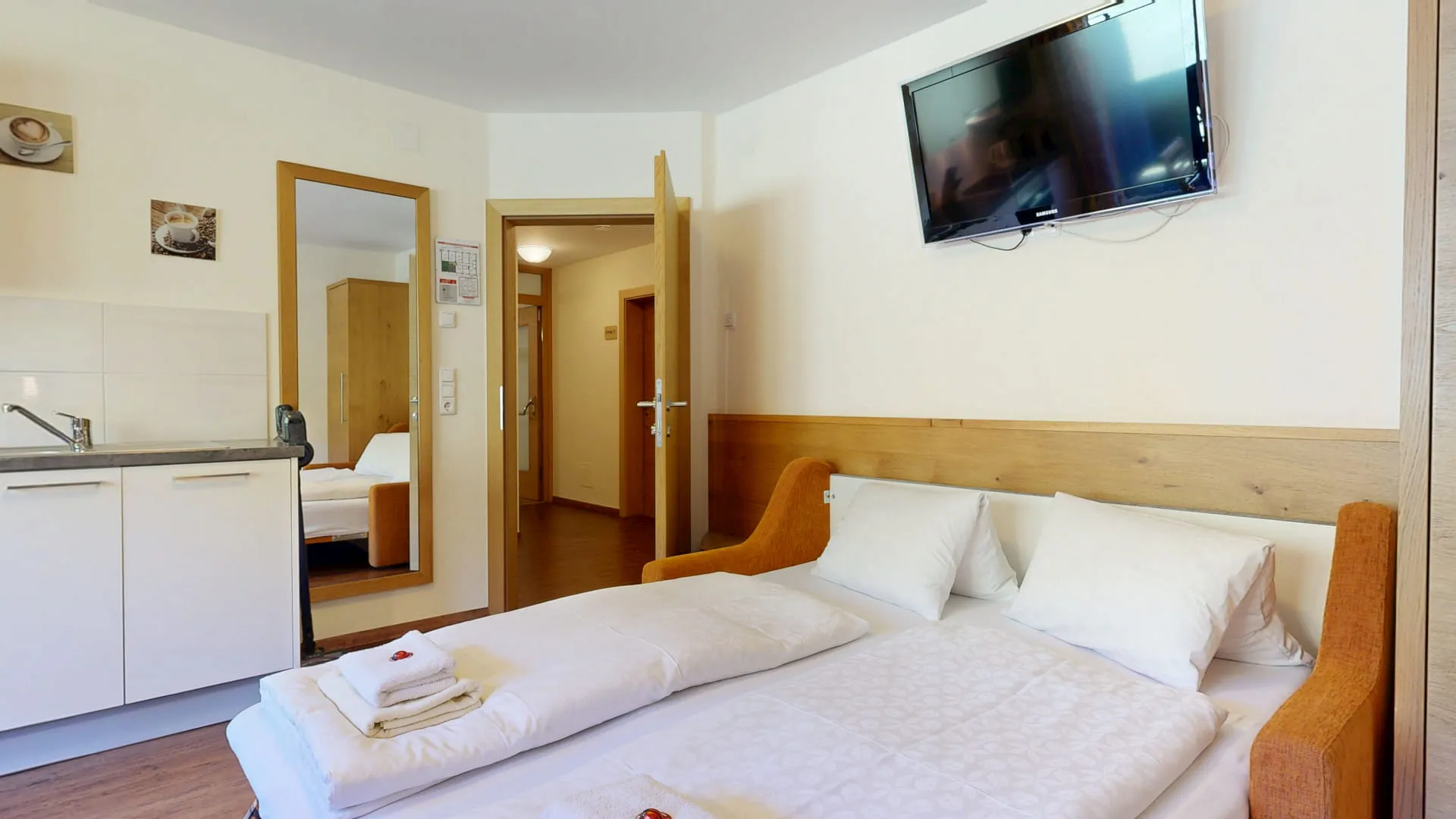 Helles Zimmer mit Doppelbett, Küchenecke und TV, Aussicht auf Flur.